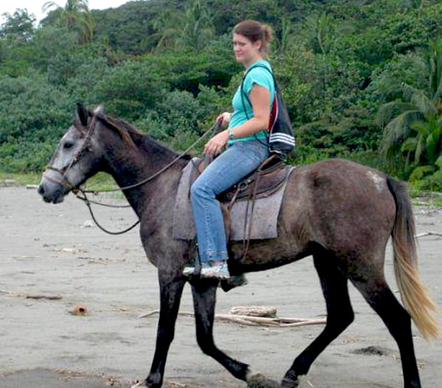 Woman riding a horse on a beach with greenery in the background